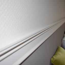 wall & floor solution - Biała listwa dekoracyjna przy suficie na tle ściany o delikatnej fakturze i szarego zagłówka łóżka z poduszkami.