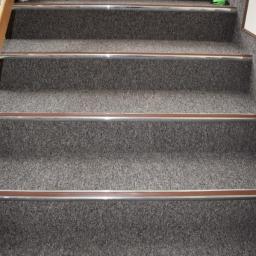 wall & floor solution - Schody pokryte szarą wykładziną z aluminiowymi listwami zabezpieczającymi krawędzie, widoczny odkurzacz w tle na górnym stopniu.