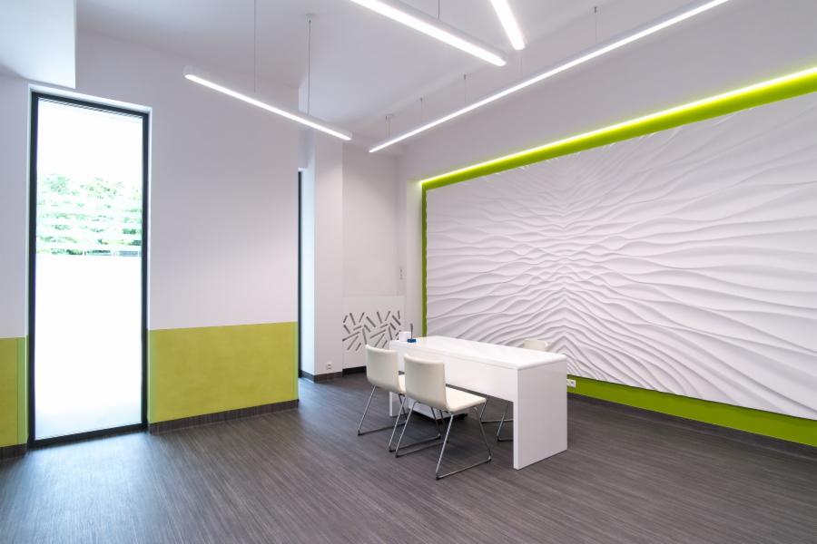 Nowoczesne biuro z białą, teksturowaną ścianą, podświetloną zielenią, szarą podłogą imitującą drewno i białymi meblami, oświetlone podłużnymi lampami LED.