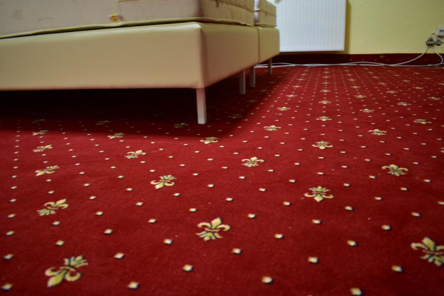 Czerwona wykładzina dywanowa w stylu retro z wzorem złotych lilii pod beżowym łóżkiem na metalowych nogach, widoczny fragment ściany z gniazdkiem elektrycznym.
