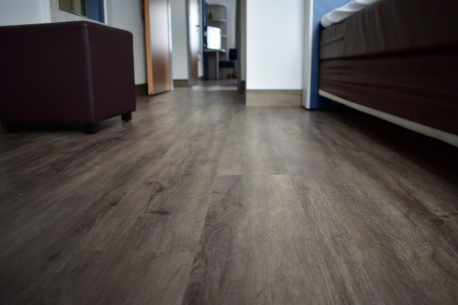 Ułożone panele podłogowe w odcieniach szarości z widoczną strukturą drewna, w tle fragment brązowego pufa i widok na dalsze pomieszczenia.