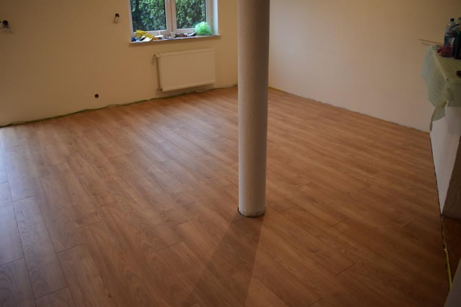Nowo ułożone panele podłogowe w pokoju z kolumną, widoczne ślady po taśmie malarskiej przy ścianach, grzejnik pod oknem.