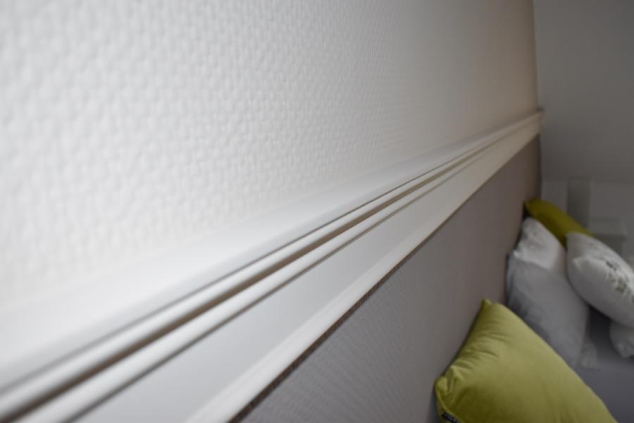 Biała listwa dekoracyjna przy suficie na tle ściany o delikatnej fakturze i szarego zagłówka łóżka z poduszkami.