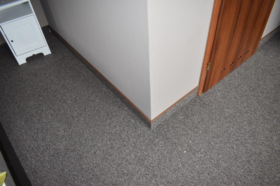 Szara wykładzina dywanowa w rogu pokoju, przy białej ścianie i drewnianej ościeżnicy drzwiowej.