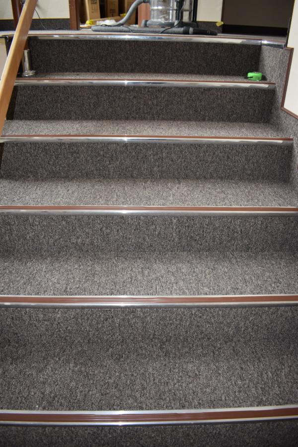Schody pokryte szarą wykładziną z aluminiowymi listwami zabezpieczającymi krawędzie, widoczny odkurzacz w tle na górnym stopniu.