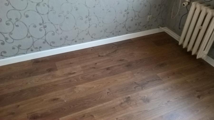Ułożone panele podłogowe w odcieniach brązu, białe listwy przypodłogowe i fragment ściany z wzorzystą tapetą w Szczecinie, widoczny stary grzejnik.