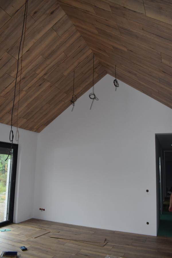 Wnętrze w trakcie prac wykończeniowych: ułożona drewnopodobna okładzina na skośnym suficie, białe ściany z widocznymi przewodami elektrycznymi, częściowo ułożona podłoga z paneli, widoczne narzędzia