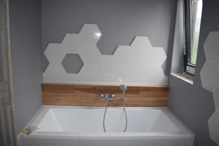 Biała wanna obudowana płytkami w kształcie heksagonów na szarej ścianie, z drewnianym panelem nad wanną i nowoczesną baterią.
