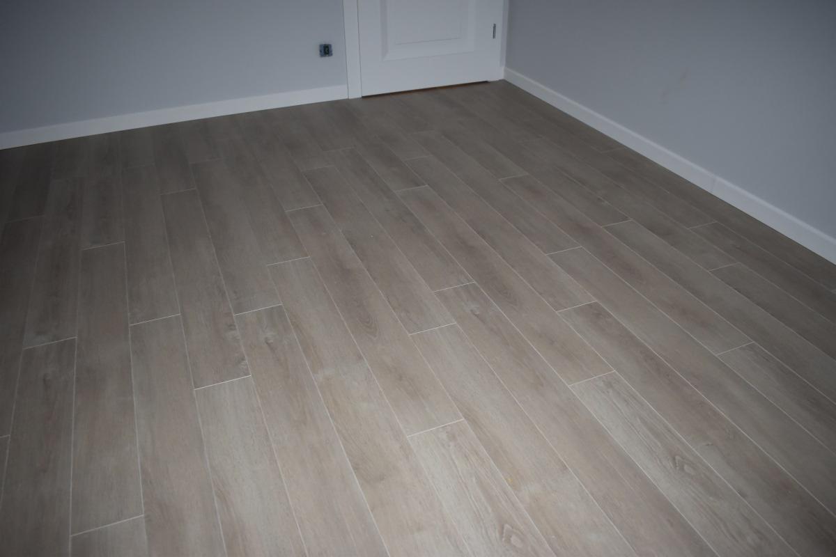 Ułożona jasna podłoga z paneli imitujących drewno, widoczne fugi i białe listwy przypodłogowe, szara ściana i białe drzwi w tle.
