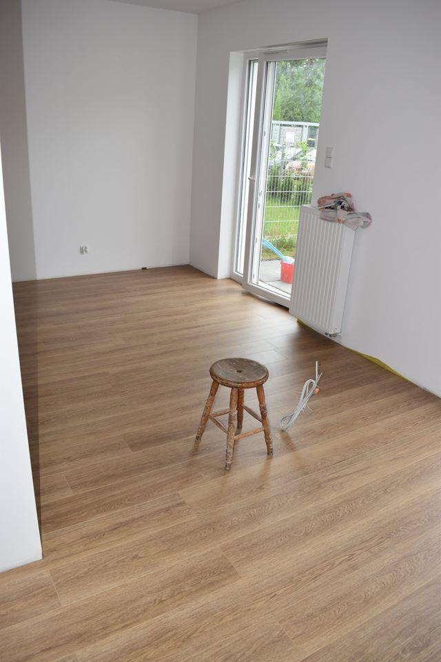 Nowo ułożone panele podłogowe o wzorze drewna w pustym pokoju z białymi ścianami, z widocznym stołkiem i wystającą instalacją elektryczną.