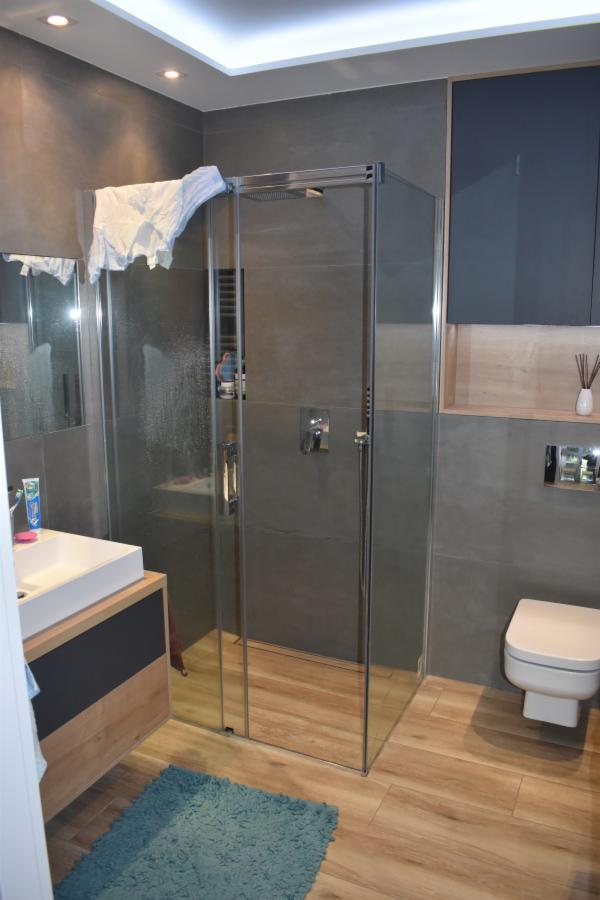 Nowoczesna łazienka z kabiną prysznicową ze szkła hartowanego, szarą glazurą, drewnopodobną podłogą i białą umywalką nablatową z szafką w odcieniach szarości i drewna, oraz turkusowym dywanikiem.