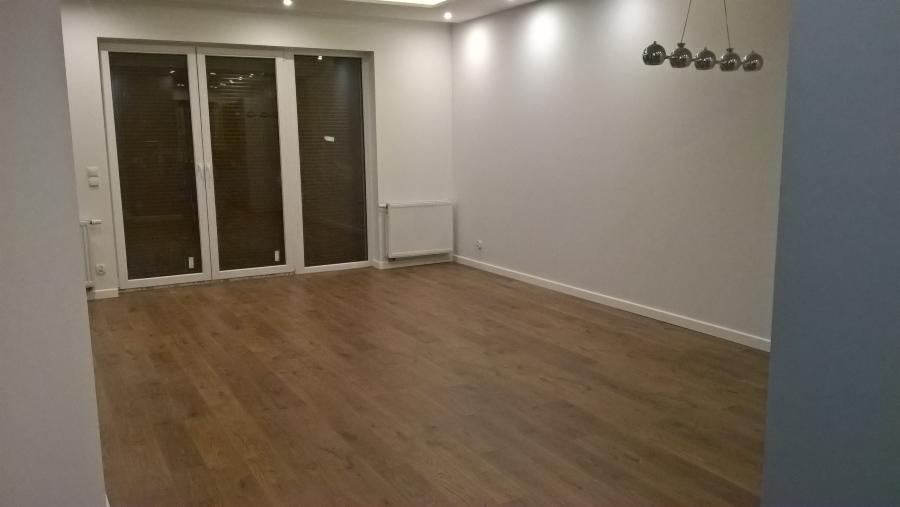 Puste, odświeżone wnętrze pokoju z drewnianą podłogą, białymi ścianami, grzejnikiem i przesuwnymi drzwiami balkonowymi z żaluzjami, oświetlone wiszącą lampą z kilkoma kloszami.