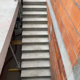 Surowe schody betonowe w trakcie budowy, widoczne szalunki i czerwona ściana z pustaków ceramicznych.