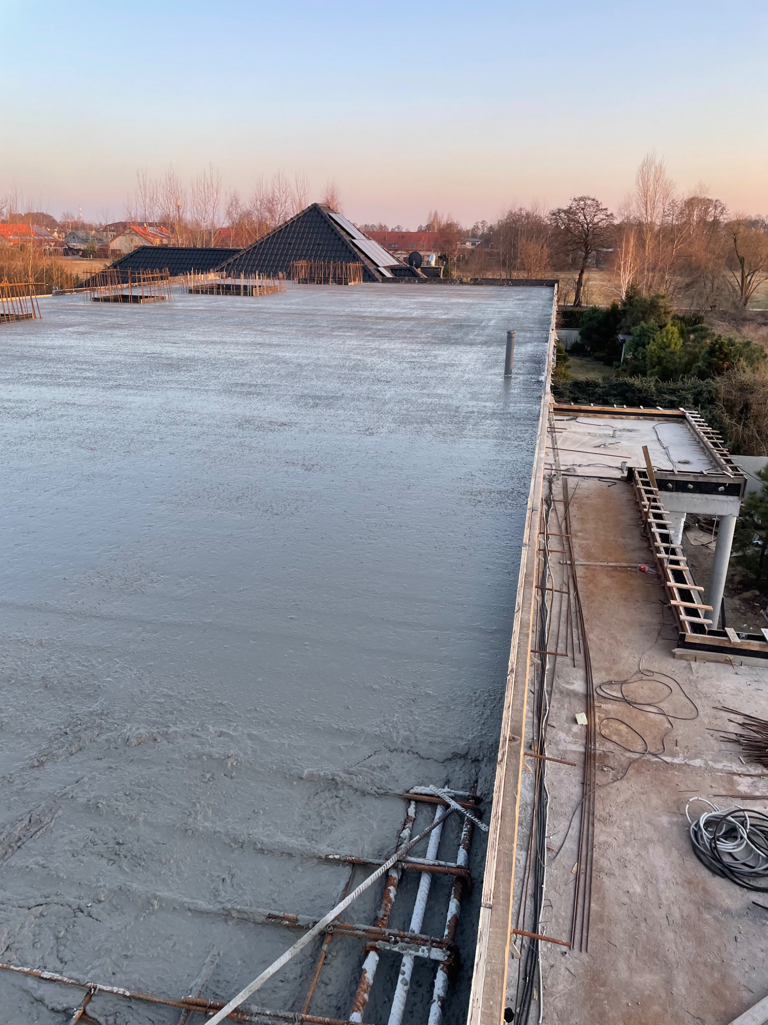 Wylany, świeży beton na stropie budynku w trakcie budowy, widoczne zbrojenia i szalunki, w tle zabudowa jednorodzinna oświetlona porannym słońcem.