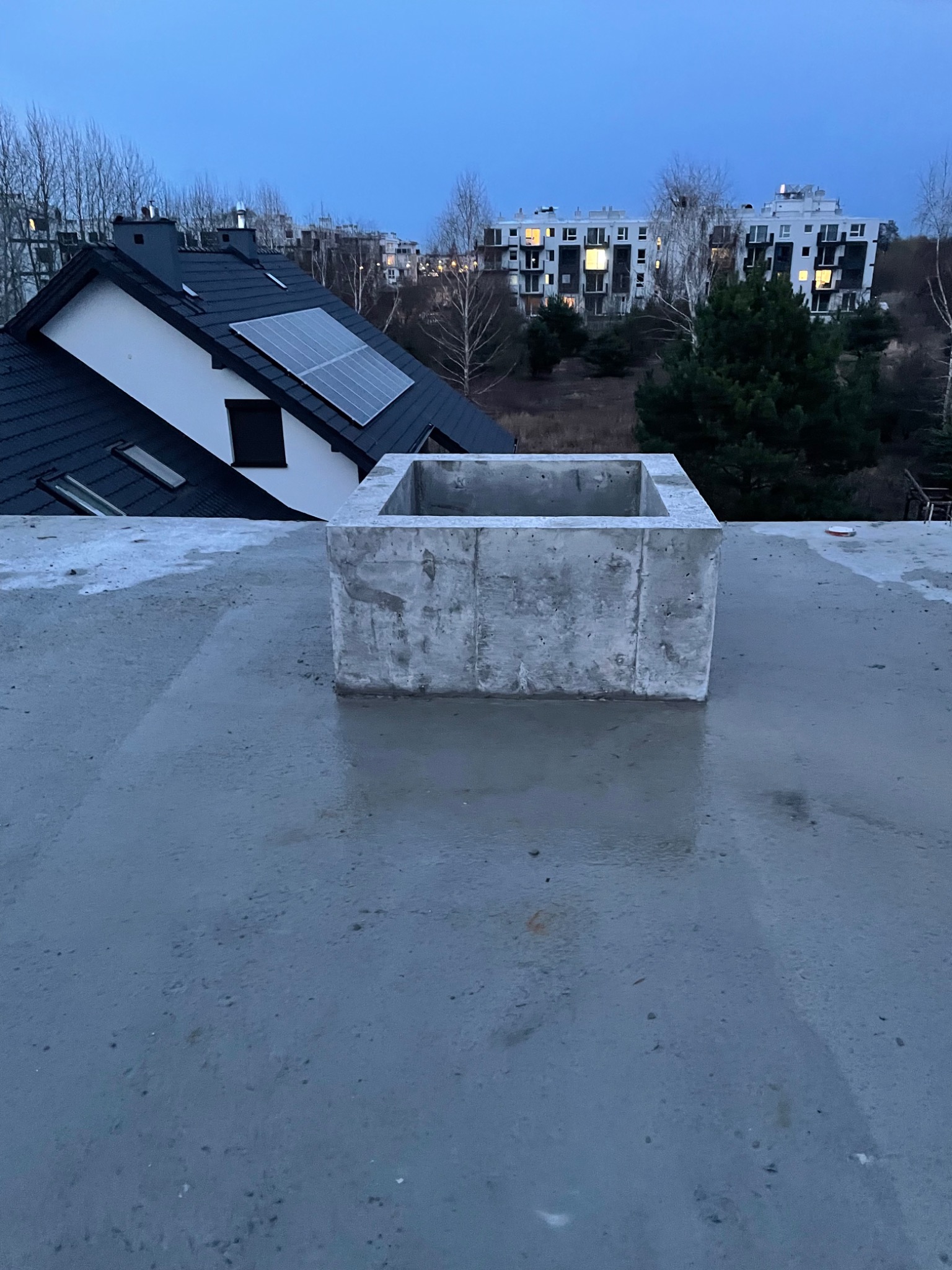 Świeżo wylany betonowy element konstrukcyjny na mokrej płycie budynku w trakcie budowy, w tle dom z panelami słonecznymi i budynki mieszkalne przy zmierzchu.