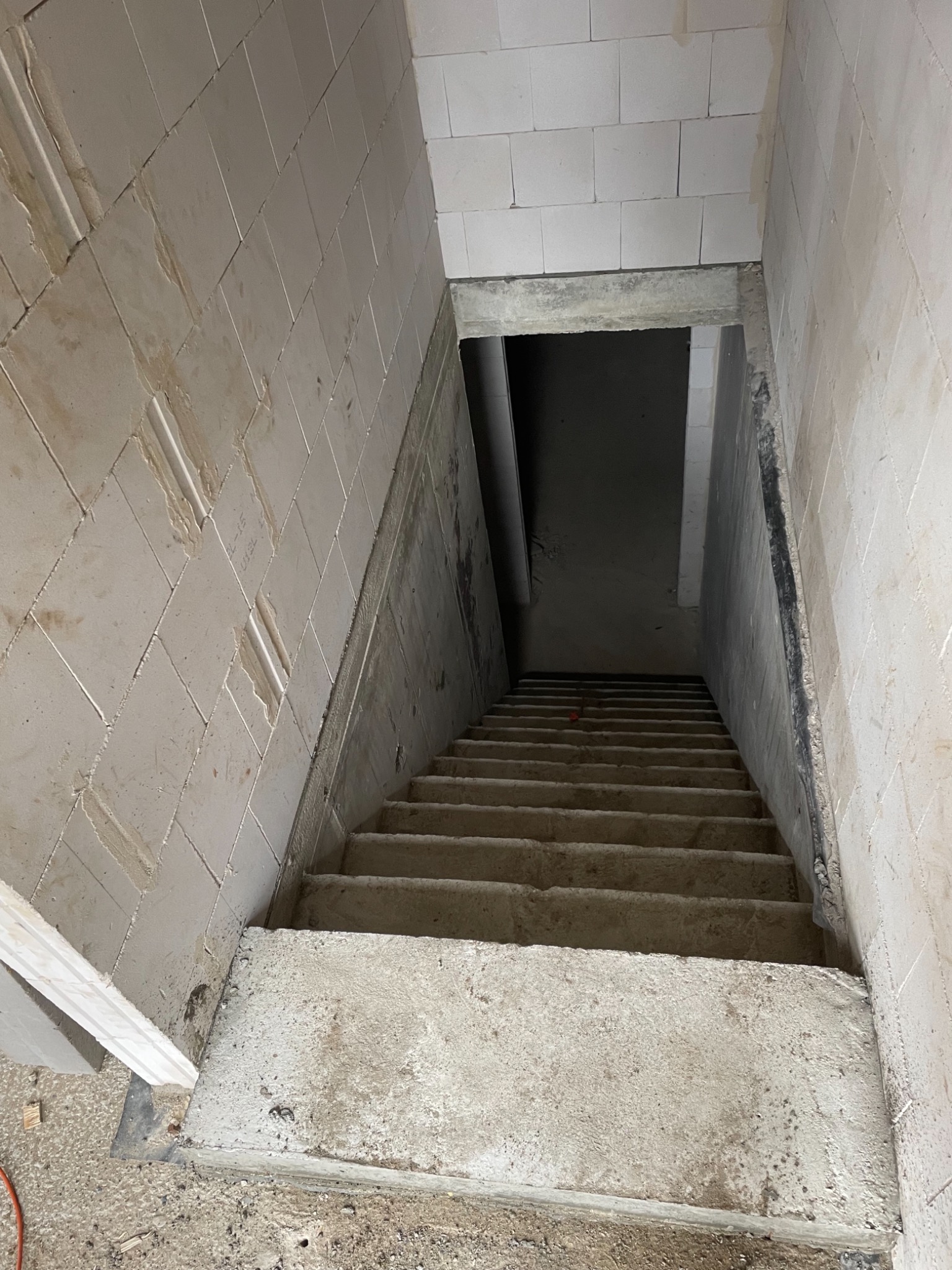 Surowe betonowe schody prowadzące w dół, z widocznymi blokami betonowymi tworzącymi ściany klatki schodowej, częściowo otynkowane, z widocznymi śladami prac budowlanych.