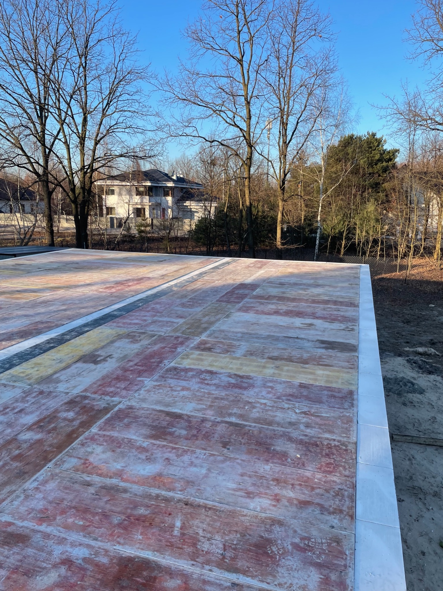 Płyta fundamentowa budynku jednorodzinnego w trakcie budowy z widocznymi warstwami izolacji i zbrojenia, na tle drzew i domu.