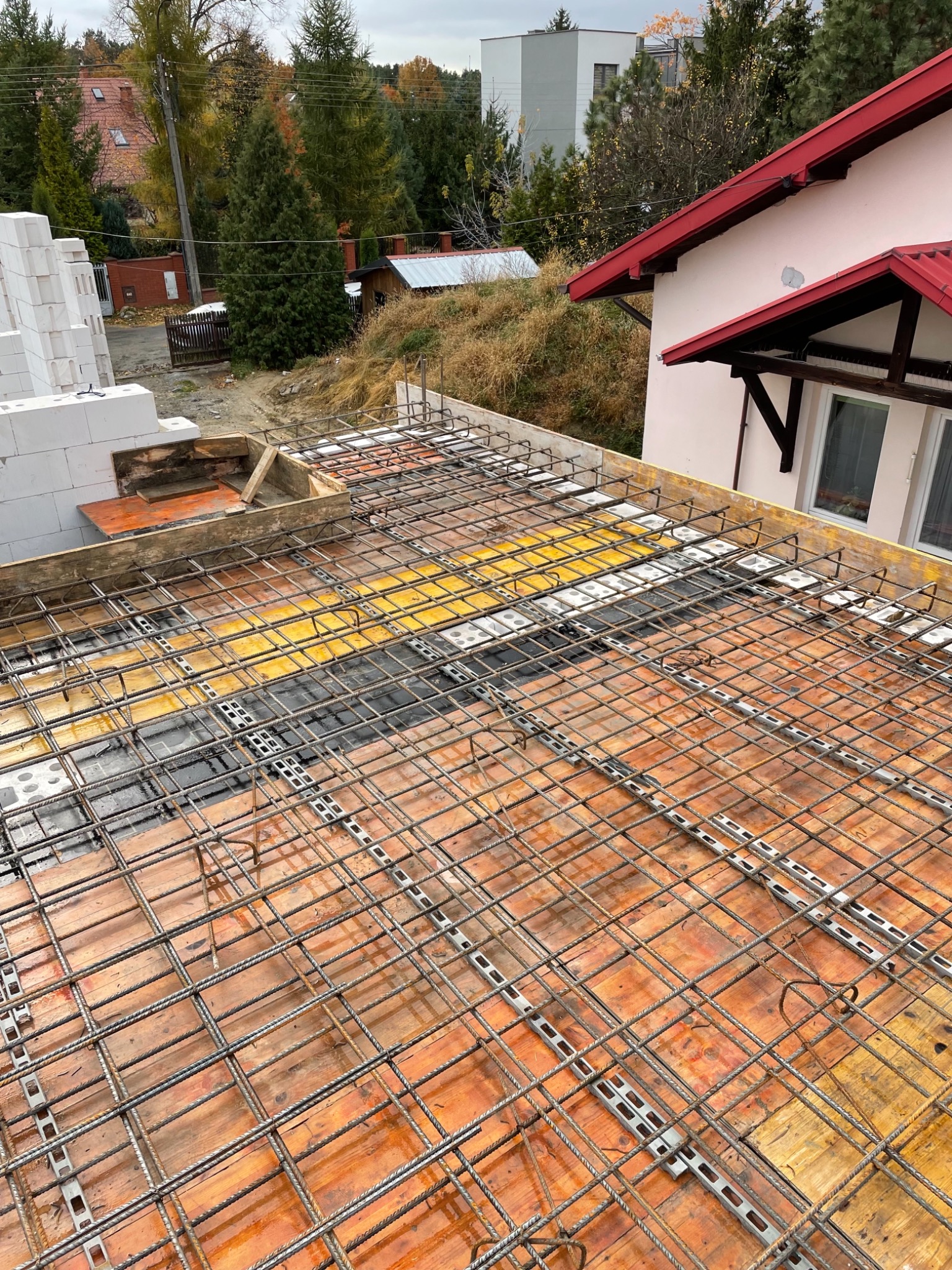 Siatka zbrojeniowa na deskowaniu stropu budynku w trakcie budowy, widoczne drewniane płyty szalunkowe, stalowe dystanse i fragment muru z bloczków gazobetonowych.