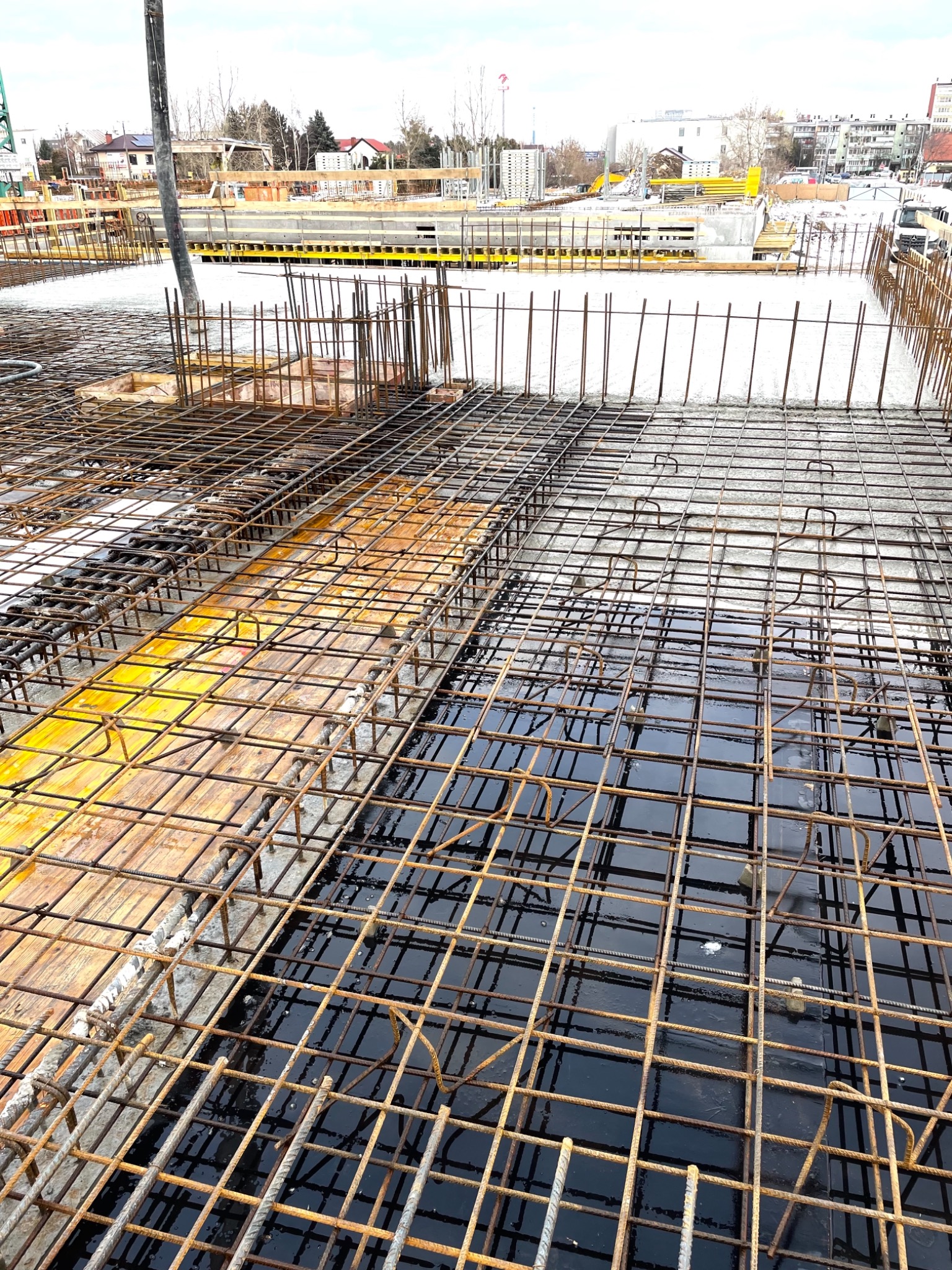 Siatka zbrojeniowa na budowie, częściowo zalana wodą, widoczne drewniane elementy szalunku i betonowe konstrukcje w tle.