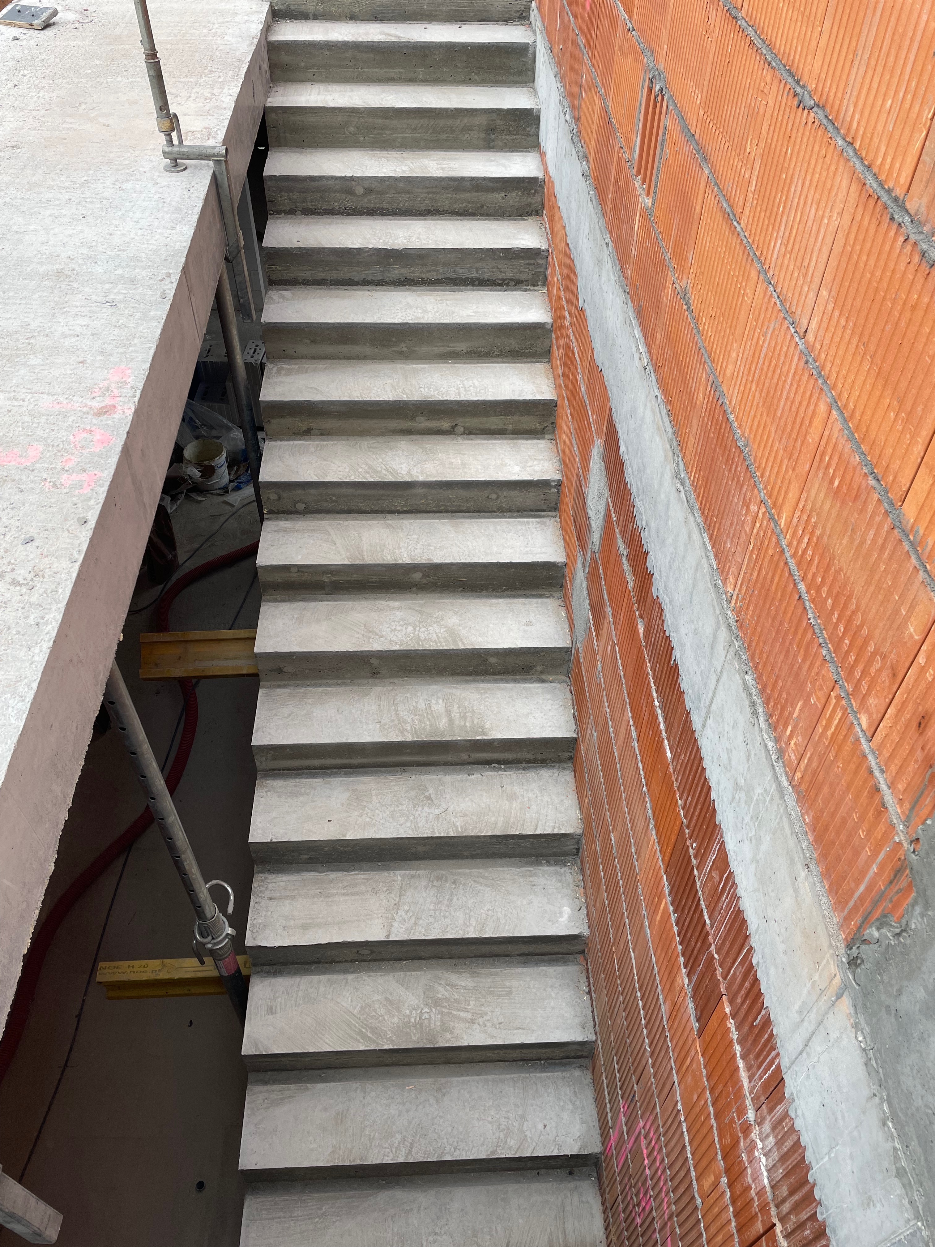 Surowe schody betonowe w trakcie budowy, widoczne szalunki i czerwona ściana z pustaków ceramicznych.