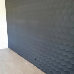 Ściana pokryta trójwymiarową tapetą w kształcie plastrów miodu, pomalowana na ciemny szary kolor, z widocznym gniazdkiem elektrycznym u dołu i jasną drewnianą podłogą.