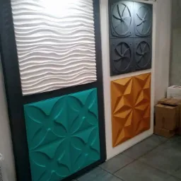 Wystawa paneli dekoracyjnych 3D: białe fale, szare wentylatory, turkusowe kwiaty i pomarańczowe origami na ścianie z oświetleniem punktowym.