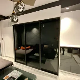 Nowoczesna szafa przesuwna z czarnymi lustrzanymi drzwiami, wkomponowana w białą zabudowę, odbijająca wnętrze sypialni z łóżkiem i lampami wiszącymi.