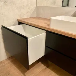 Czarna szafka łazienkowa z wysuwaną szufladą, drewniany blat i prostokątna umywalka nablatowa na tle jasnych płytek.