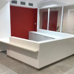 Biała lada recepcyjna z szafkami w tle, ściana pomalowana na czerwono, widoczne drzwi i okno.