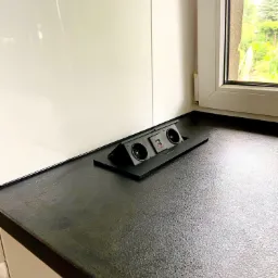 Czarny blat kuchenny z wbudowanym, wysuwanym gniazdkiem elektrycznym i portami USB, widok z boku, w tle okno z widokiem na zieleń.