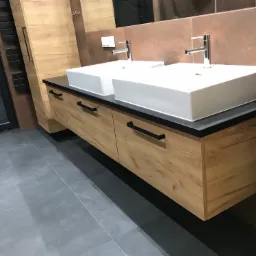 Nowoczesna łazienka z dwiema prostokątnymi umywalkami nablatowymi, osadzonymi na szafce z drewnopodobnymi frontami i czarnym blatem, uzupełniona o minimalistyczne baterie i brązowe płytki na ścianie.