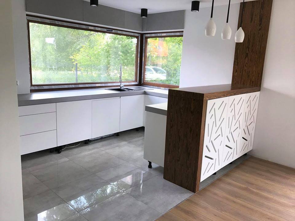 Nowoczesna kuchnia z białymi szafkami i szarym blatem pod dużym oknem, z elementami dekoracyjnymi z ciemnego drewna i białymi, ażurowymi frontami szafek, oraz trzema wiszącymi lampami.