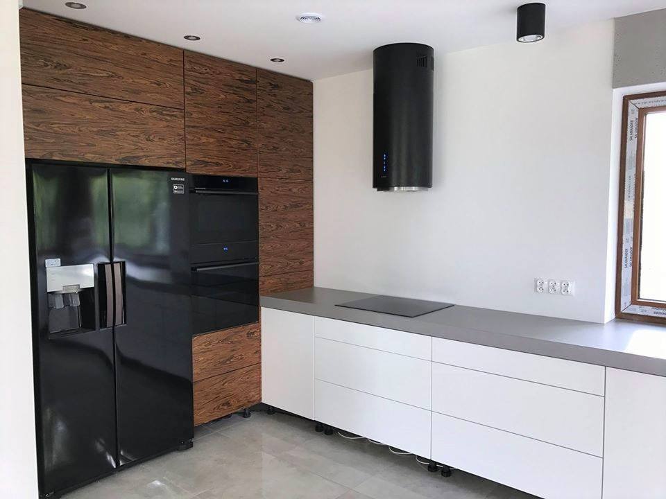 Nowoczesna kuchnia z białymi szafkami dolnymi, drewnianymi szafkami górnymi, czarną lodówką Samsung i cylindrycznym okapem, oświetlona punktowymi lampami sufitowymi.