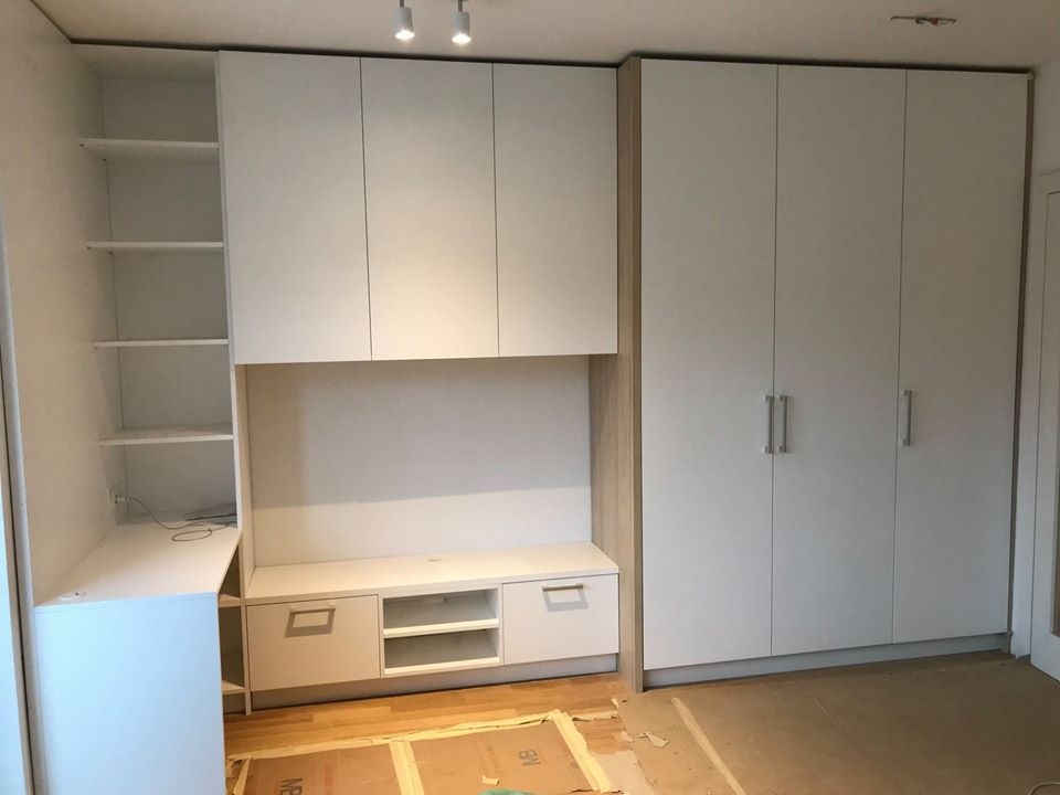 Białe meble na wymiar w zabudowie: szafa z czterema drzwiami, szafki wiszące i regał z półkami, widoczne kartony na podłodze podczas montażu.
