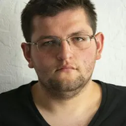 Portret mężczyzny w okularach na jasnym tle. Ujęcie od przodu, skupione na twarzy, z delikatnym cieniem po prawej stronie. Wyraz twarzy neutralny, włosy krótkie.