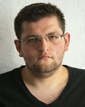 Portret mężczyzny w okularach na jasnym tle. Ujęcie od przodu, skupione na twarzy, z delikatnym cieniem po prawej stronie. Wyraz twarzy neutralny, włosy krótkie.