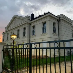 Elegancki, dwupiętrowy dom w stylu klasycystycznym z kremową elewacją, widoczny zza ozdobnego, metalowego ogrodzenia i oświetlony latarnią uliczną o zmierzchu.