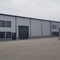 Nowo wybudowana hala magazynowa z szarej blachy trapezowej, widoczne okna, drzwi i brama wjazdowa, na dachu drabina ewakuacyjna, teren wokół utwardzony kostką brukową.