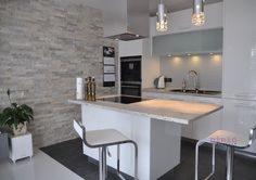 Nowoczesna kuchnia z wyspą w bieli i szarości. Ściana wykończona dekoracyjną płytką imitującą kamień, oświetlenie punktowe nad wyspą i blatem, minimalistyczne krzesła barowe z białymi siedziskami.