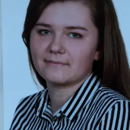 Portret młodej kobiety z długimi brązowymi włosami, ubranej w czarno-białą koszulę w paski, na jasnym tle.