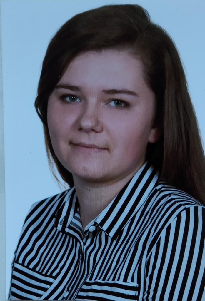 Portret młodej kobiety z długimi brązowymi włosami, ubranej w czarno-białą koszulę w paski, na jasnym tle.