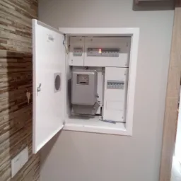 Otwarta skrzynka elektryczna z licznikiem i bezpiecznikami na ścianie z dekoracyjną okładziną, widoczny klucz w zamku i wkrętak wewnątrz.