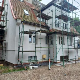 Dom jednorodzinny w trakcie wykonywania elewacji, otoczony rusztowaniem, z widoczną częściowo odnowioną dachówką i nowym tynkiem na ścianach.