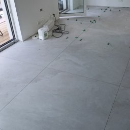 AMBER-BUD PAWEŁ ŚLIWIŃSKI - Układanie jasnych, kwadratowych płytek podłogowych z użyciem zielonych krzyżyków dystansowych w Wiślince, widoczne przygotowane materiały i przewody elektryczne.