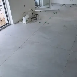 Układanie jasnych, kwadratowych płytek podłogowych z użyciem zielonych krzyżyków dystansowych w Wiślince, widoczne przygotowane materiały i przewody elektryczne.