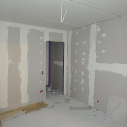 AMBER-BUD PAWEŁ ŚLIWIŃSKI - Wnętrze w trakcie prac wykończeniowych: ściany pokryte płytami kartonowo-gipsowymi z widocznymi otworami po wkrętach, surowa podłoga, wystające przewody elektryczne i fragmenty kartonów na podłodze...