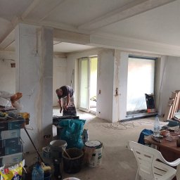 AMBER-BUD PAWEŁ ŚLIWIŃSKI - Wnętrze w trakcie prac wykończeniowych, widoczny mężczyzna szlifujący ścianę, liczne narzędzia i materiały budowlane, surowe ściany z widocznymi śladami po elektryce, listwy dekoracyjne pod sufitem...