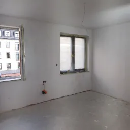 Pok&oacute;j w trakcie prac wykończeniowych: ściany po nałożeniu gładzi, zabezpieczone okna folią malarską, widoczne puszki elektryczne i wystające przewody, surowa podłoga.