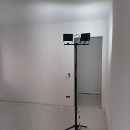 AMBER-BUD PAWEŁ ŚLIWIŃSKI - Wnętrze w trakcie malowania ścian na biało, widoczny statyw z dwoma lampami halogenowymi oświetlającymi pomieszczenie, surowy beton na podłodze, ściany po gładziowaniu, przygotowane do dalszych...