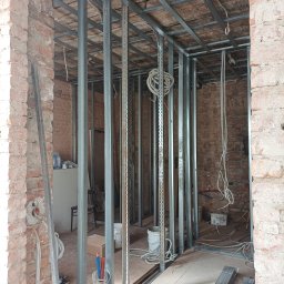 AMBER-BUD PAWEŁ ŚLIWIŃSKI - Stelaż z profili metalowych zamontowany w pomieszczeniu z surową, czerwoną cegłą, widoczne instalacje elektryczne i hydrauliczne, przygotowanie do montażu płyt gipsowo-kartonowych.